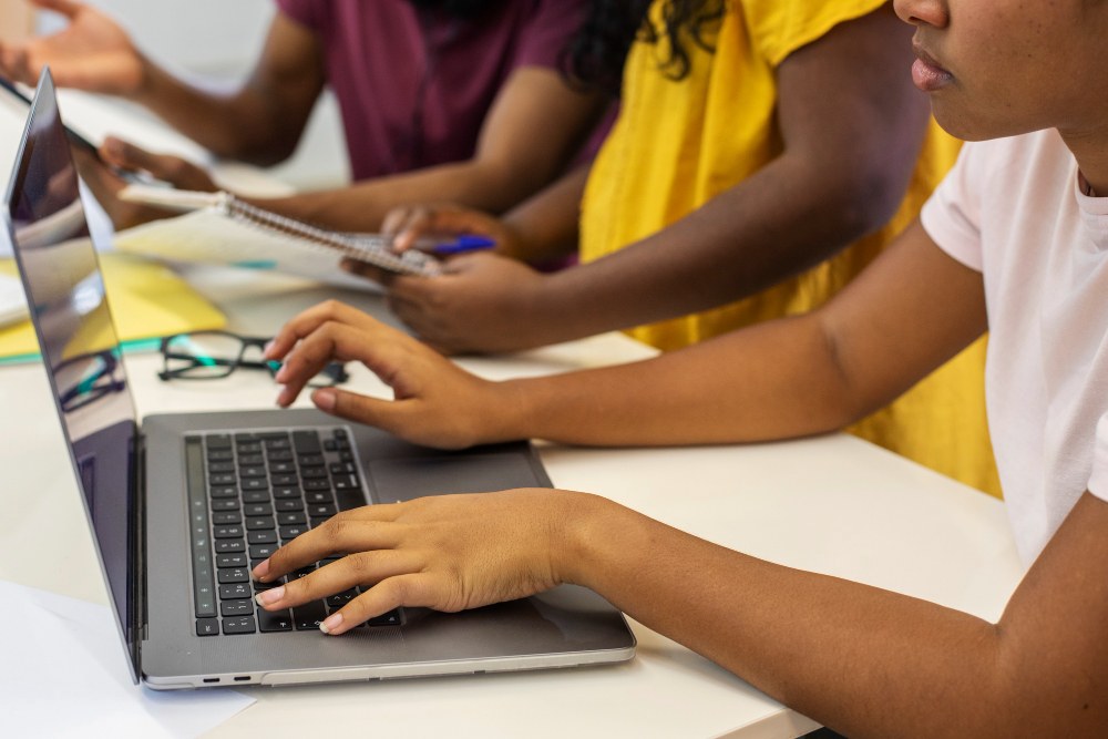 Mulher digitando em um Notebook enquanto seus colegas no funcho escrevem em cadernos.