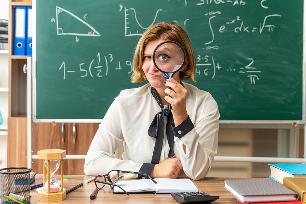 Professora de matemática em sua mesa segurando uma lupa e fazendo careta.