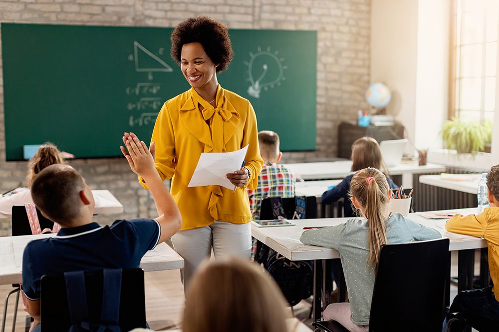 Professora em pé na sala de aula cumprimentando aluno que está sentado.