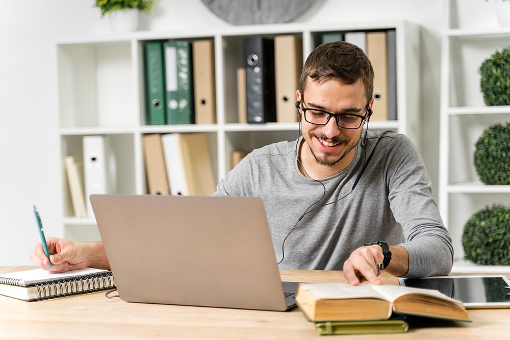 Estudante de óculos usando fone de ouvido enquanto lê livros e faz anotações.