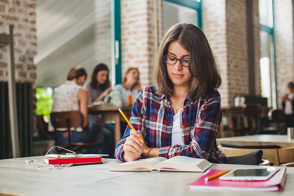 Estudante em biblioteca fazendo redação e lendo livros.