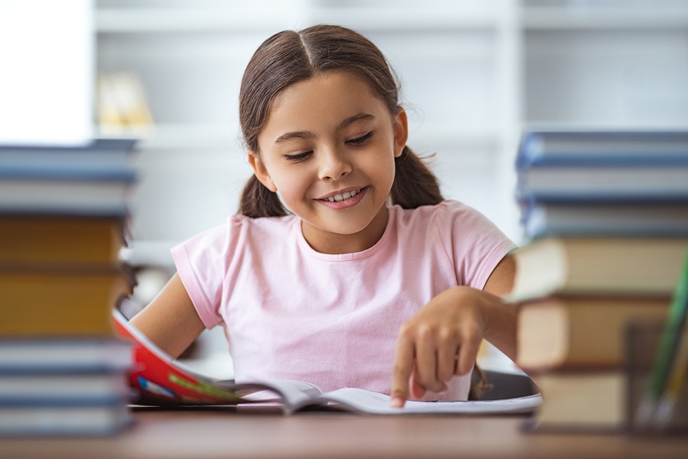 Menina em sala de aula lendo livro em voz alta ao lado de uma pilha de livros.