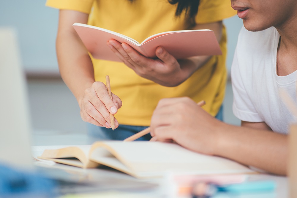 Professora em pé ao lado de um aluno do ensino médio tirando dúvidas.